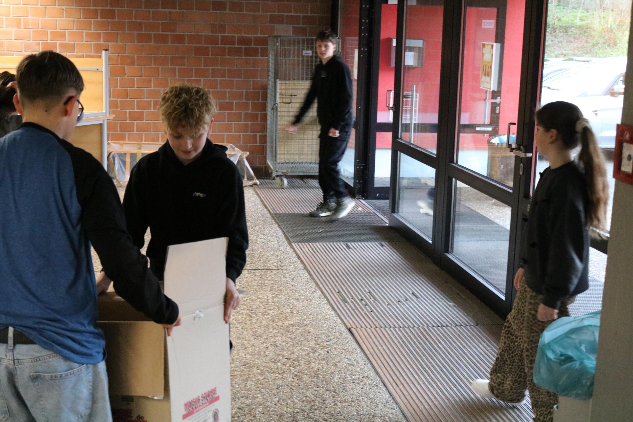 Schüler:innen tragen im Eingangsbereich der Schule große Kartons für die Weihnachtskistenaktion. Zwei Kinder heben gemeinsam einen Karton, ein weiteres Kind kommt durch die Tür hinein, eine Schülerin steht daneben und beobachtet den Vorgang.