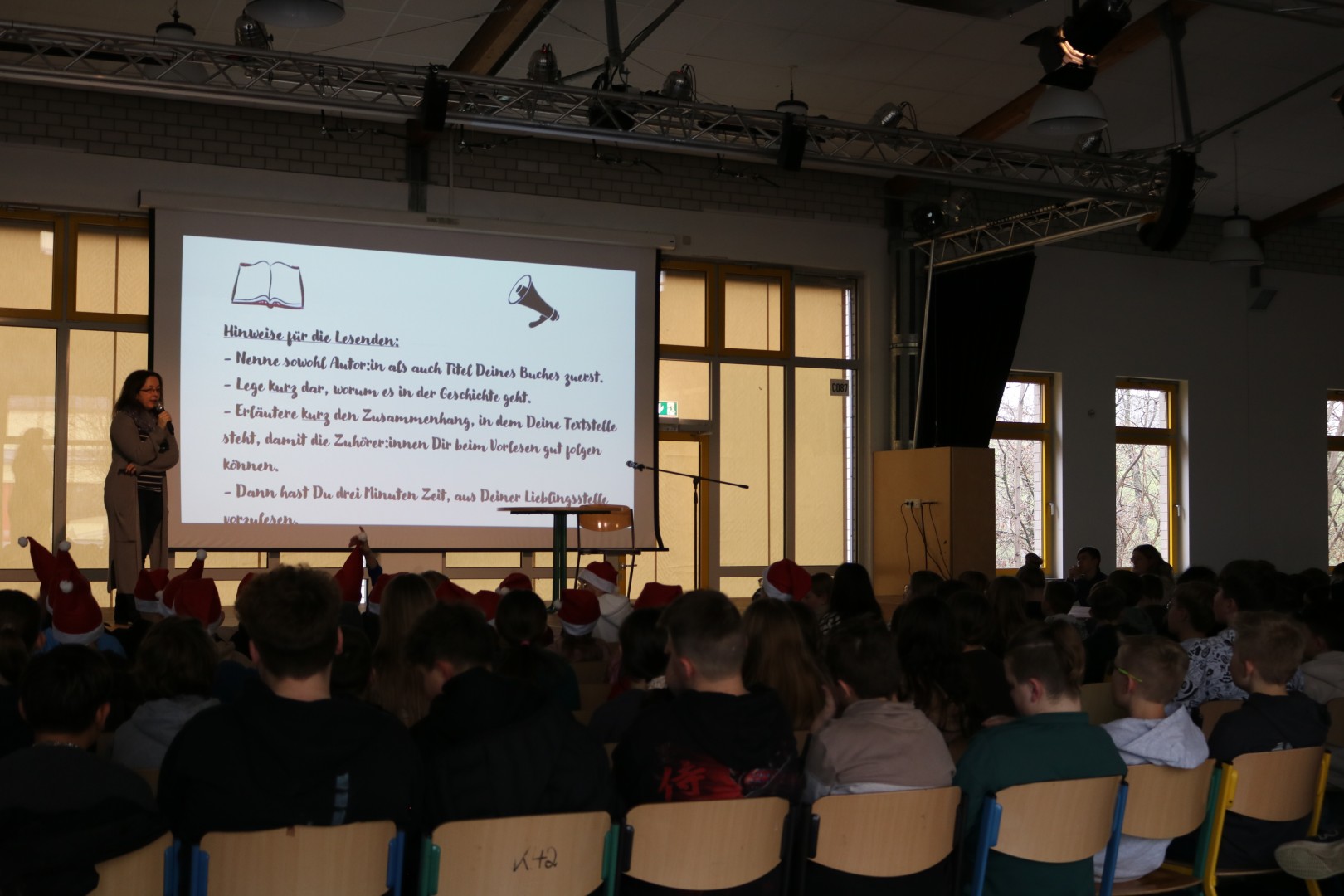 Blick von hinten auf das sitzende Publikum in der Mensa; auf der Bühne ist eine Projektionsfläche mit Hinweisen für die Vorleser:innen zu sehen.