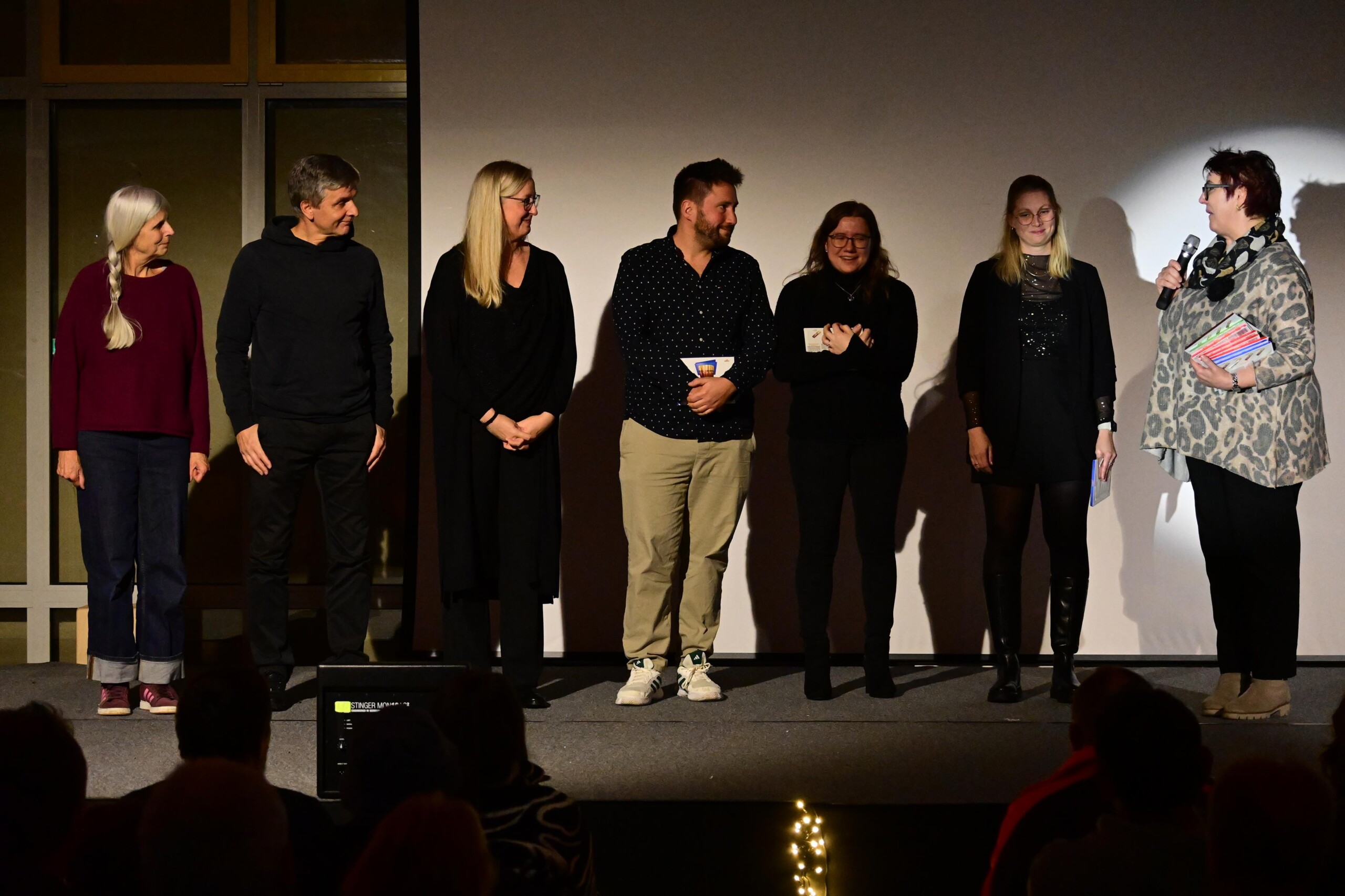 Lehrkräfte und Organisator:innen stehen auf der Bühne, eine Person spricht mit Mikrofon, die anderen hören zu, Publikum im Vordergrund sichtbar.
