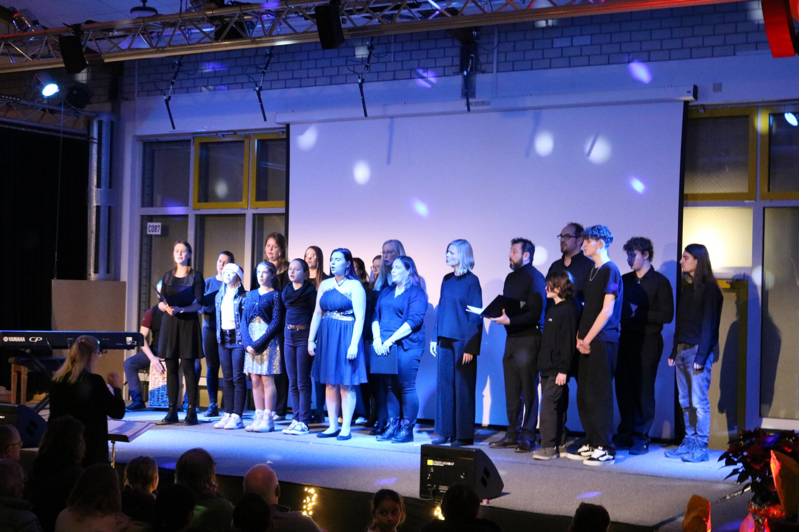 Schüler:innen- und Lehrer:innen-Chor steht gemeinsam auf der Bühne, einige halten Notenmappen, links begleitet eine Person am Keyboard, Publikum im Vordergrund sichtbar.