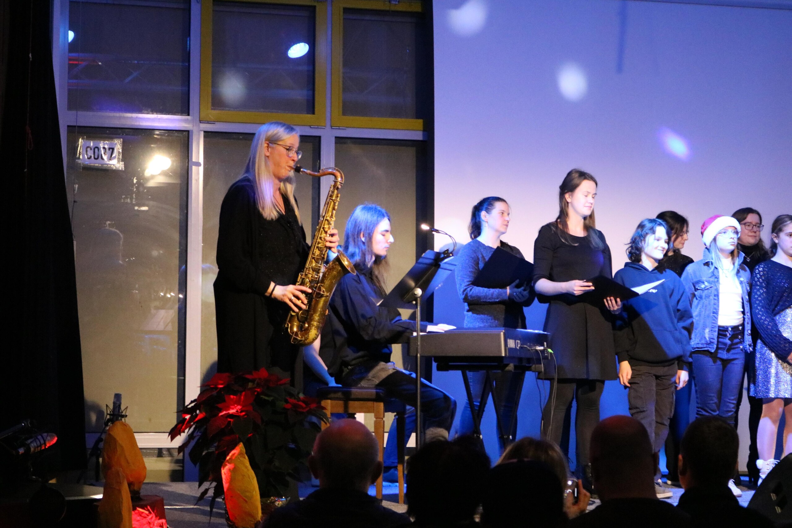 Lehrerin spielt Saxophon auf der Bühne, daneben sitzt ein Schüler am Keyboard, weitere Chormitglieder stehen im Hintergrund unter blauem Bühnenlicht.