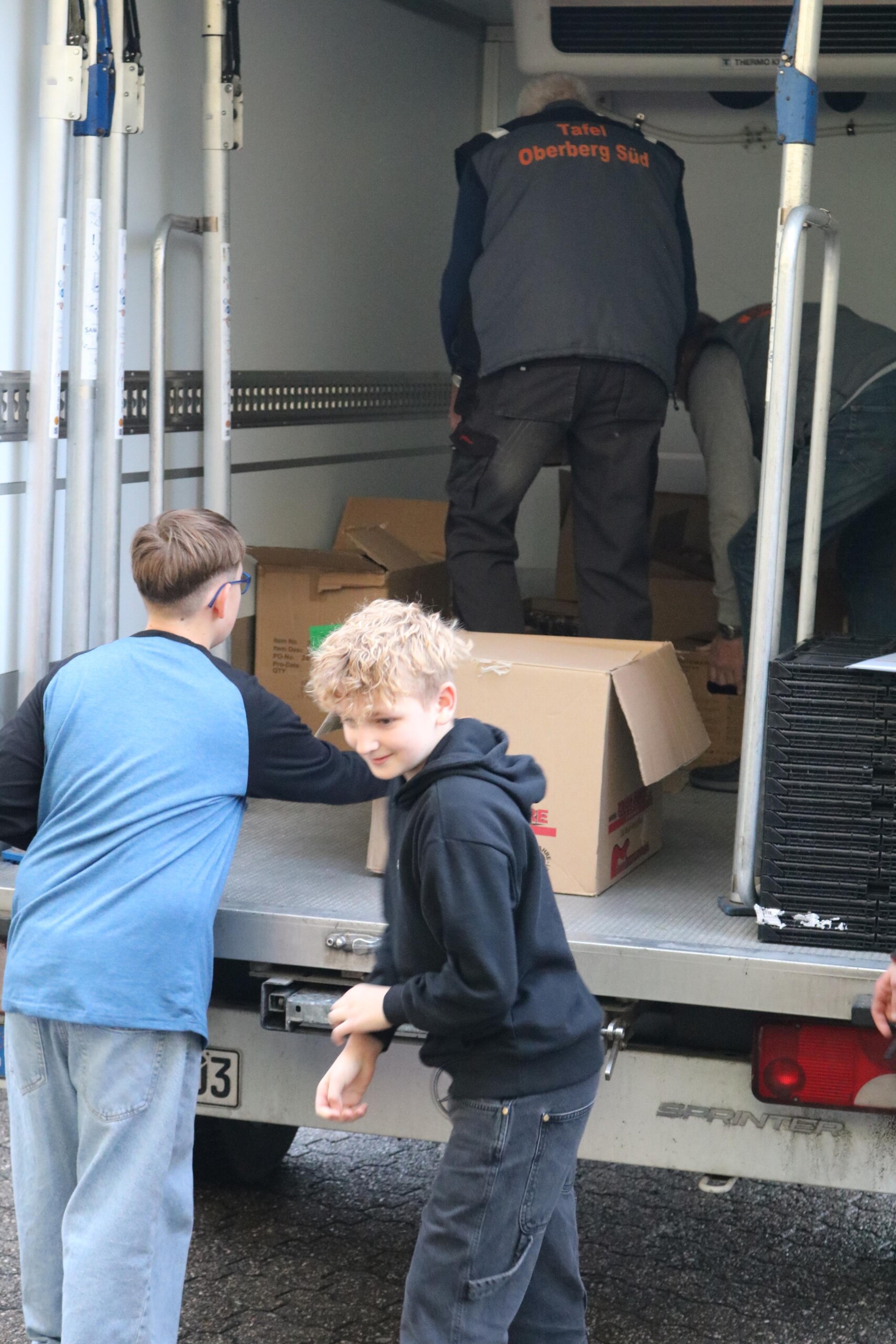Schüler:innen laden Kartons in den Laderaum eines Lieferwagens der Tafel Oberberg Süd. Ein Kind reicht einen Karton nach oben, während zwei Mitarbeitende der Tafel im Wageninneren weitere Kisten sortieren.
