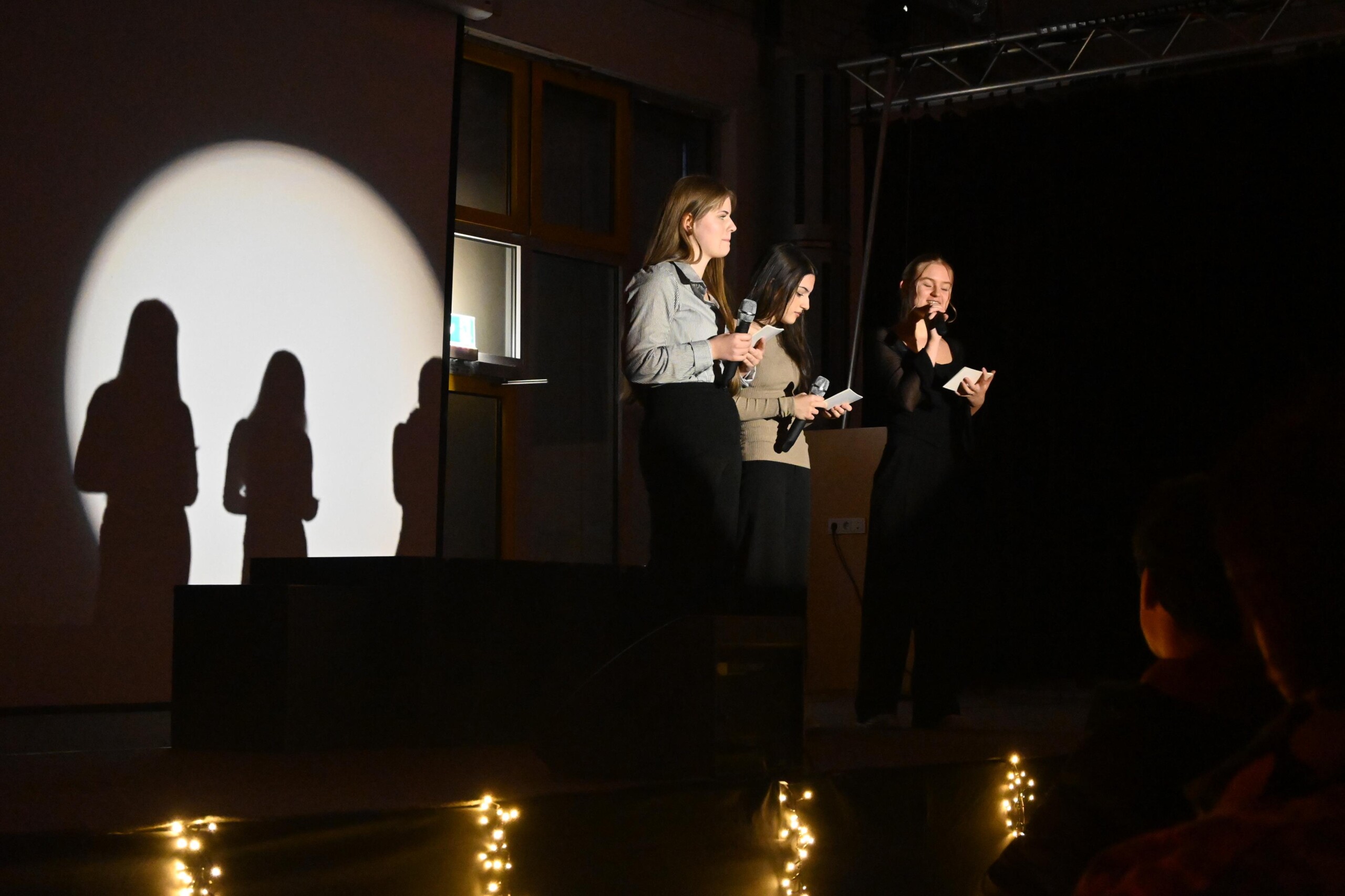 Drei Schülerinnen moderieren die Veranstaltung „Lichter im Advent“ auf der Bühne, mit Mikrofonen in der Hand und Beleuchtung im Hintergrund.
