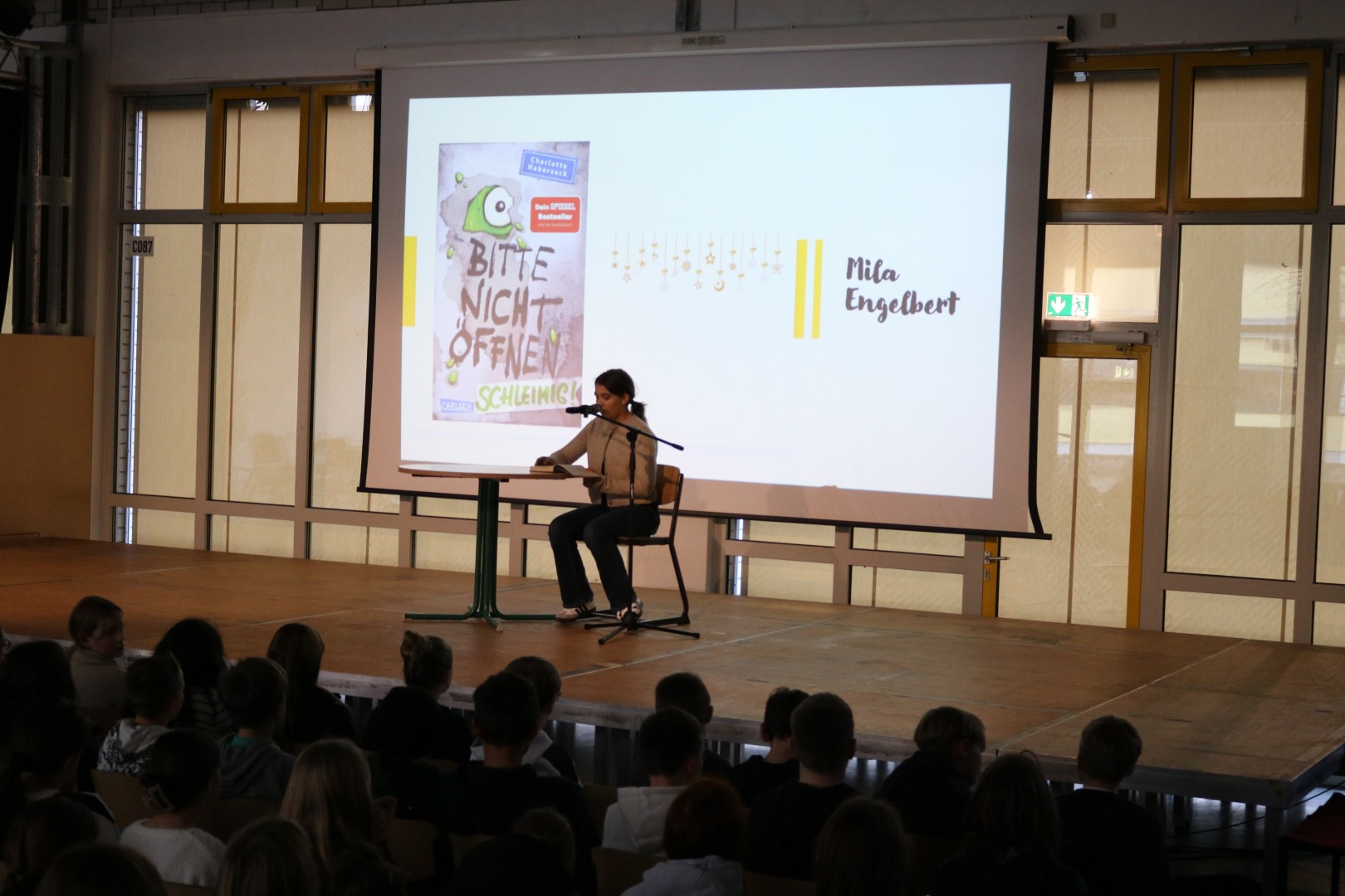 Schülerin sitzt an einem Tisch auf der Bühne und liest aus einem Buch vor; auf der Leinwand ist das Buchcover „Bitte nicht öffnen. Schleimig“ zu sehen.