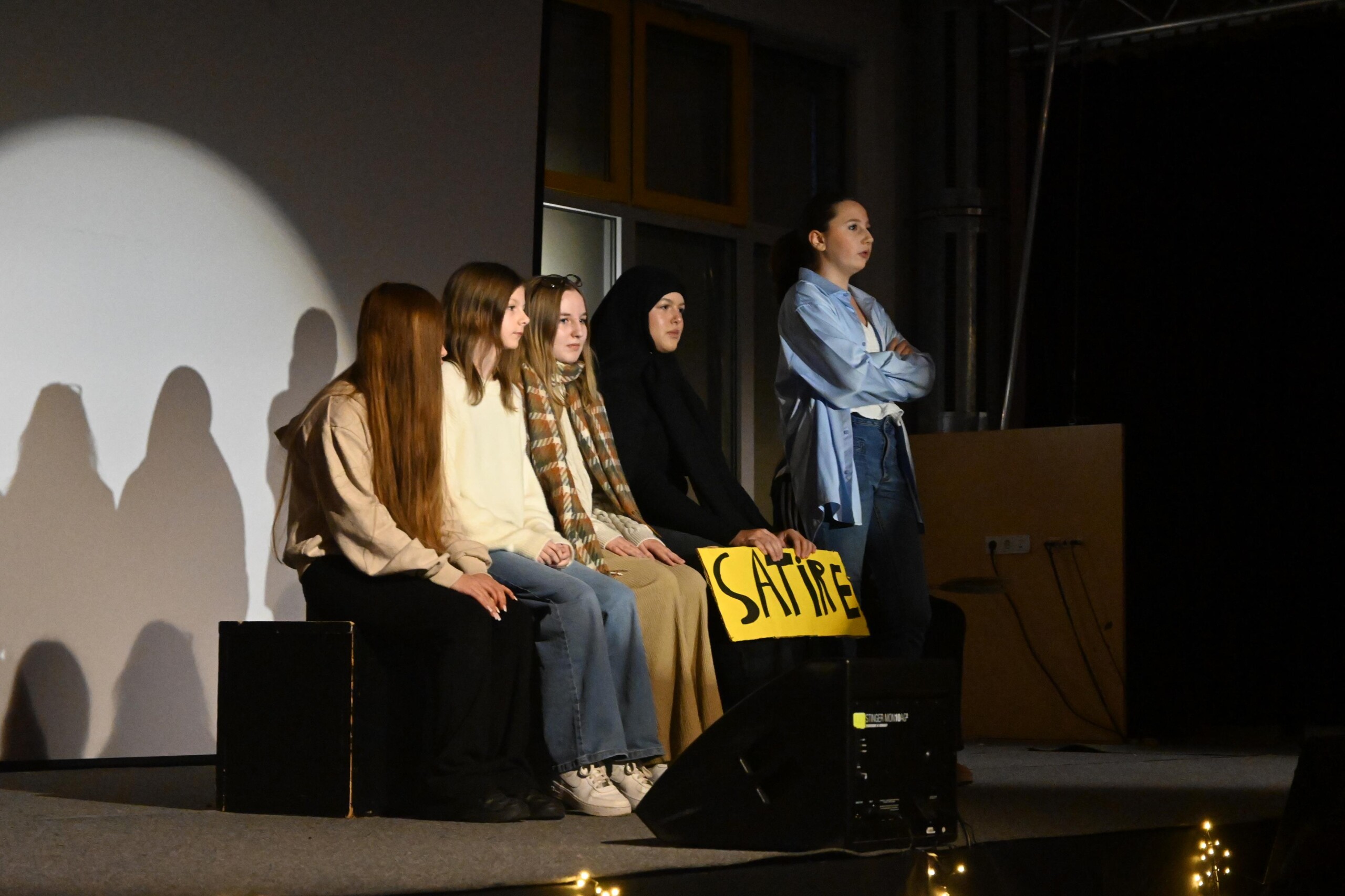 Szenische Darstellung des Profilkurses Darstellung und Gestalten: mehrere Schüler:innen sitzen und stehen auf der Bühne, eine Schülerin spricht zum Publikum, Requisit mit der Aufschrift „Satire“.