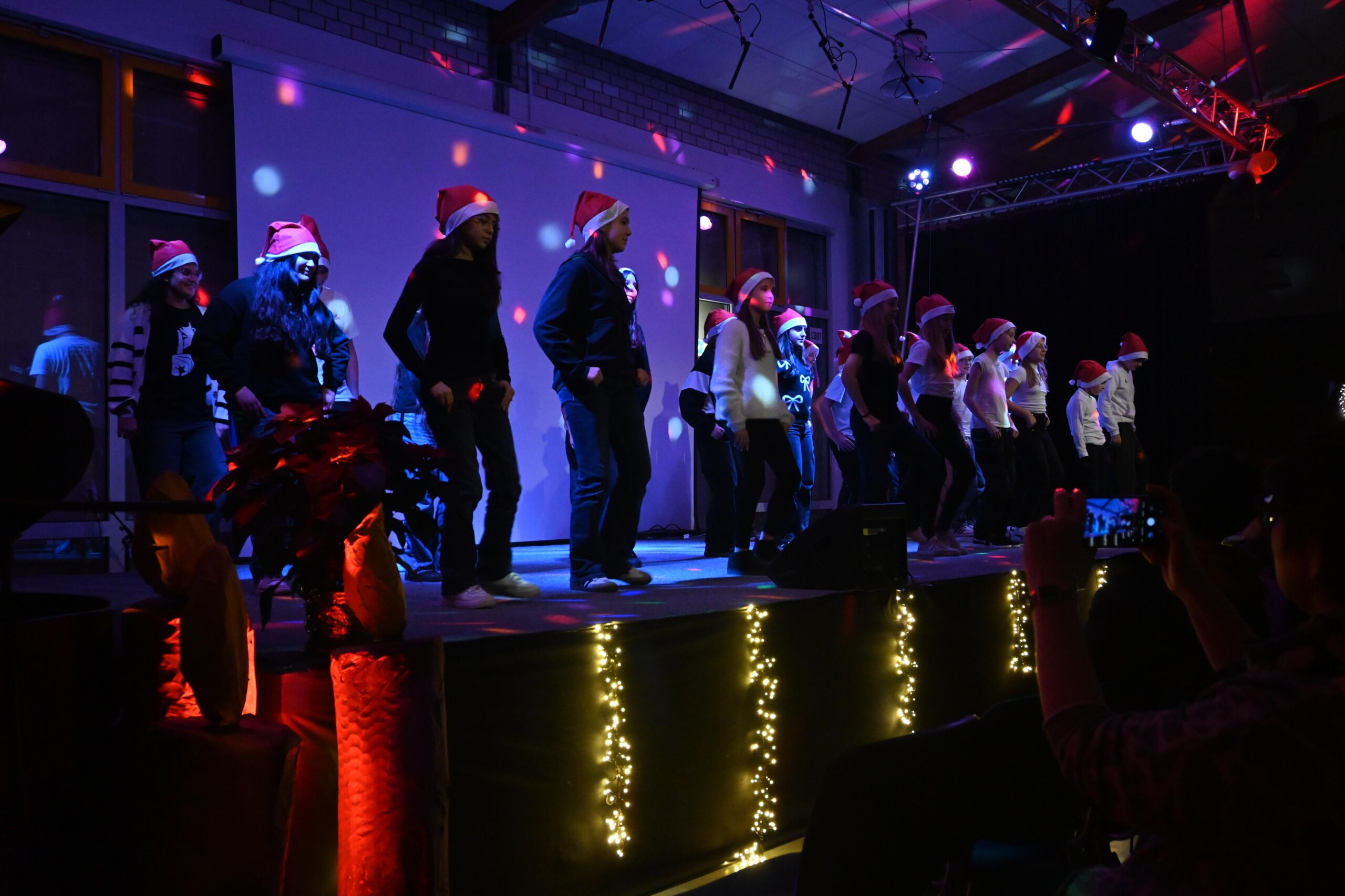 Schüler:innen der Klasse 8c tanzen gemeinsam auf der Bühne, viele tragen Nikolausmützen, beleuchtet von farbigem Bühnenlicht bei „Lichter im Advent“.