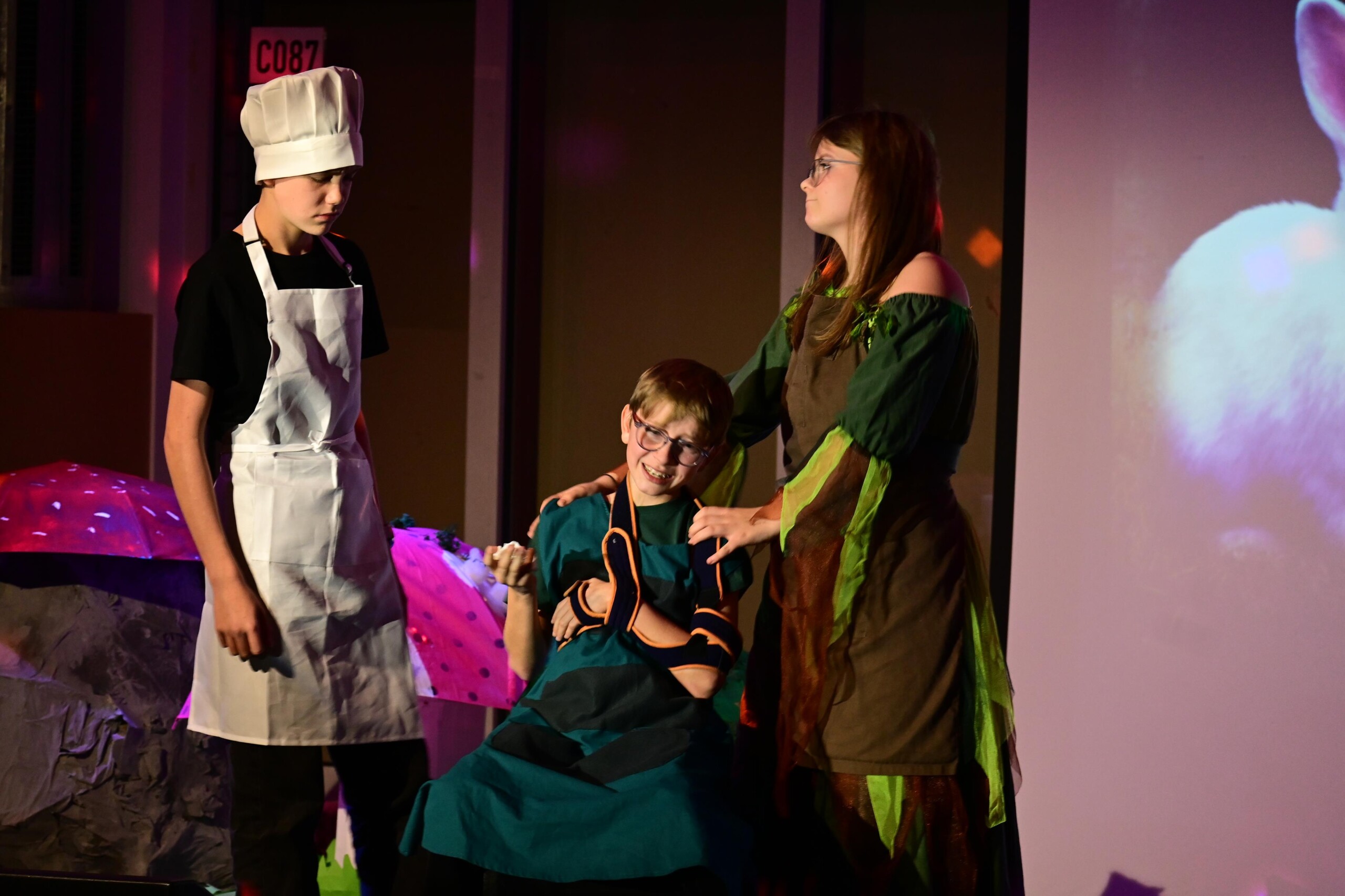 Szenenbild aus "Alice im Wunderland": drei Schüler:innen in Kostümen stehen auf der Bühne, eine Person sitzt im Rollstuhl als Teil der Darstellung.