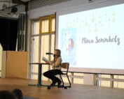 Schülerin liest an einem Stehtisch aus einem Buch; auf der Leinwand ist „Pferdeflüsterer Mädchen – Rubys Entscheidung“ zu sehen.