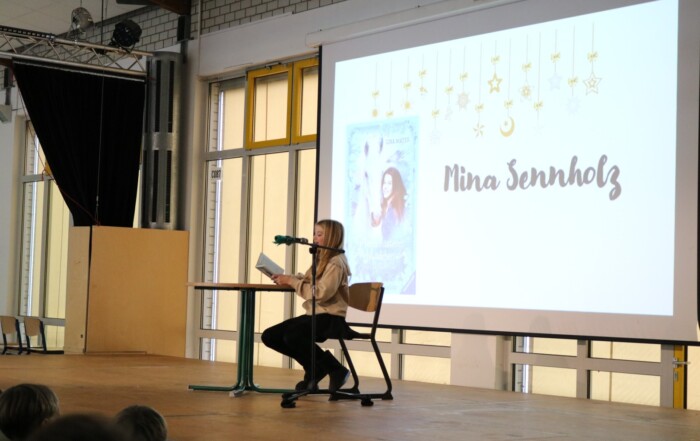 Schülerin liest an einem Stehtisch aus einem Buch; auf der Leinwand ist „Pferdeflüsterer Mädchen – Rubys Entscheidung“ zu sehen.