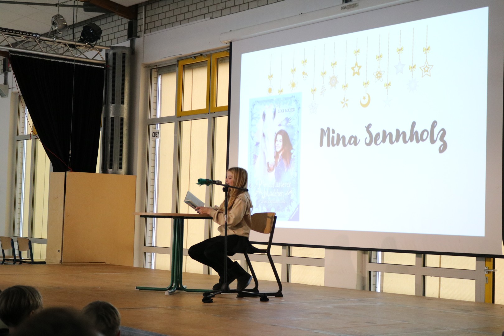 Schülerin liest an einem Stehtisch aus einem Buch; auf der Leinwand ist „Pferdeflüsterer Mädchen – Rubys Entscheidung“ zu sehen.