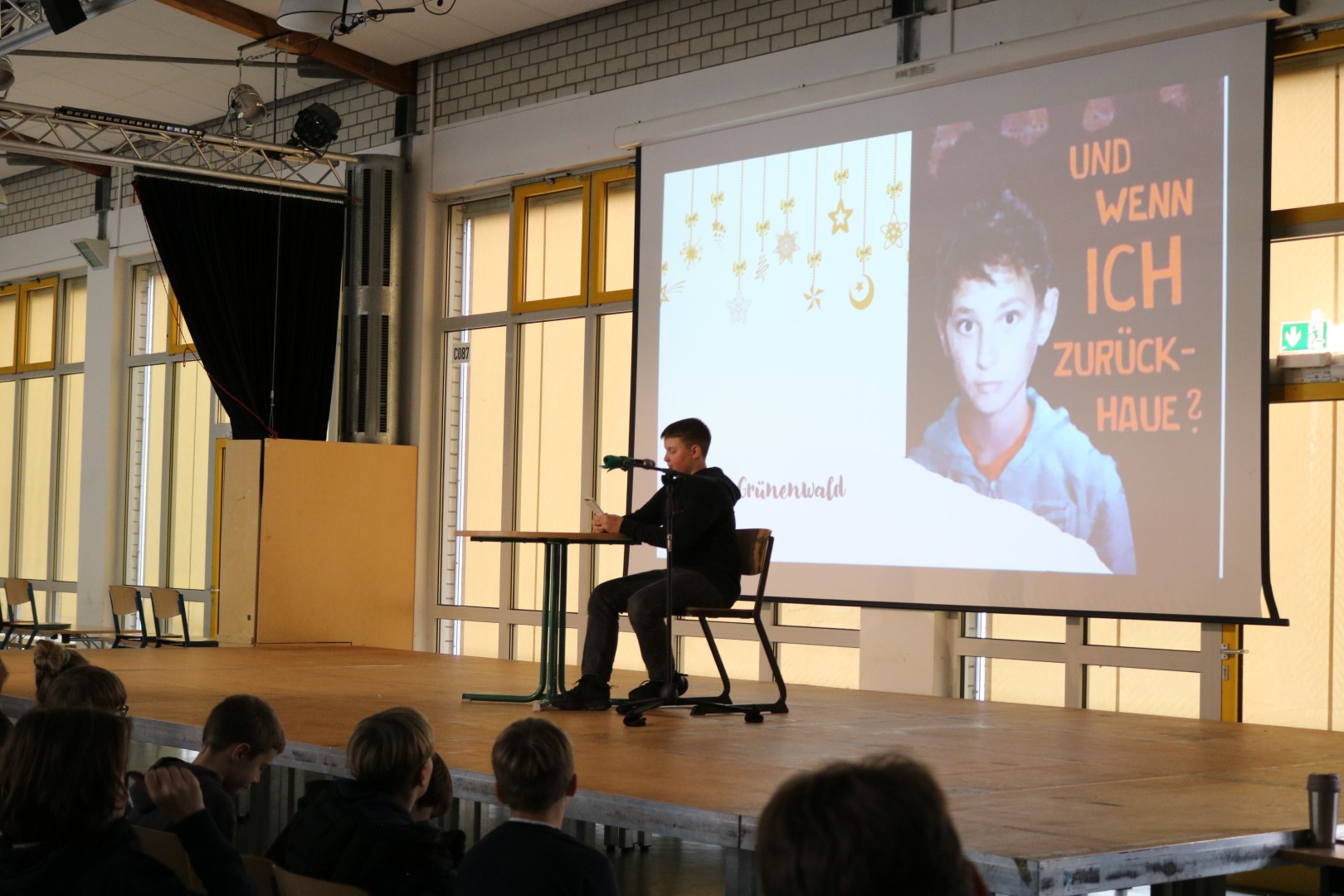 Schüler sitzt auf der Bühne und liest; auf der Leinwand ist das Buch „Und wenn ich zurückhaue?“ zu erkennen.