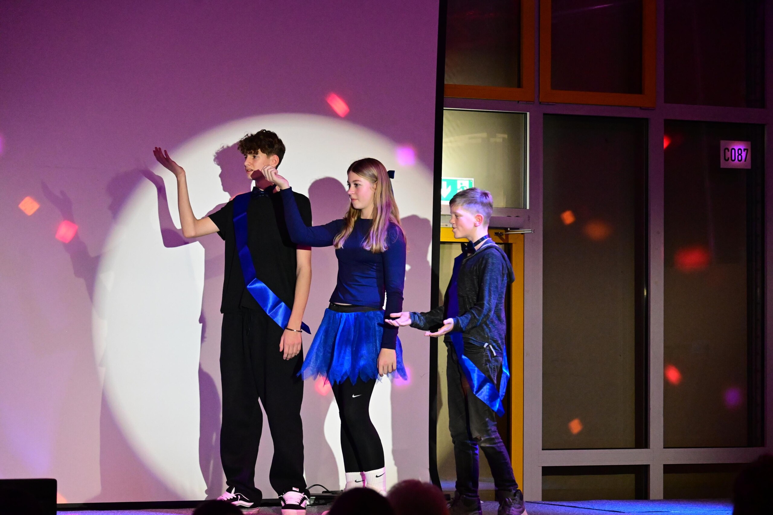 Szenische Darstellung: drei Schüler:innen stehen vor einer Projektionsfläche, eine Schülerin spricht, zwei weitere stehen seitlich.