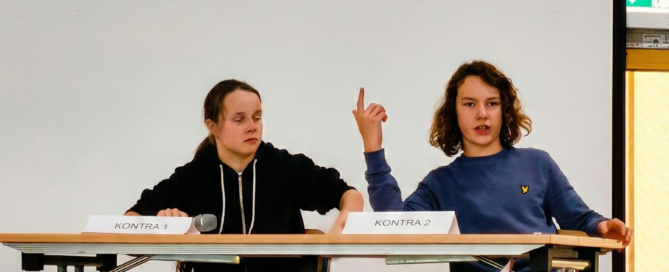 Collage aus drei Bildern einer Debattenveranstaltung auf einer Bühne vor weißem Hintergrund. Das obere Bild zeigt die Gesamtszene mit zwei Zweierteams an Tischen; ein:e Teilnehmer:in auf der rechten Seite spricht gestikulierend in ein Mikrofon. Die unteren Detailaufnahmen zeigen das Pro-Team beim konzentrierten Mitschreiben sowie das Kontra-Team während eines Redebeitrags mit erhobenem Zeigefinger.