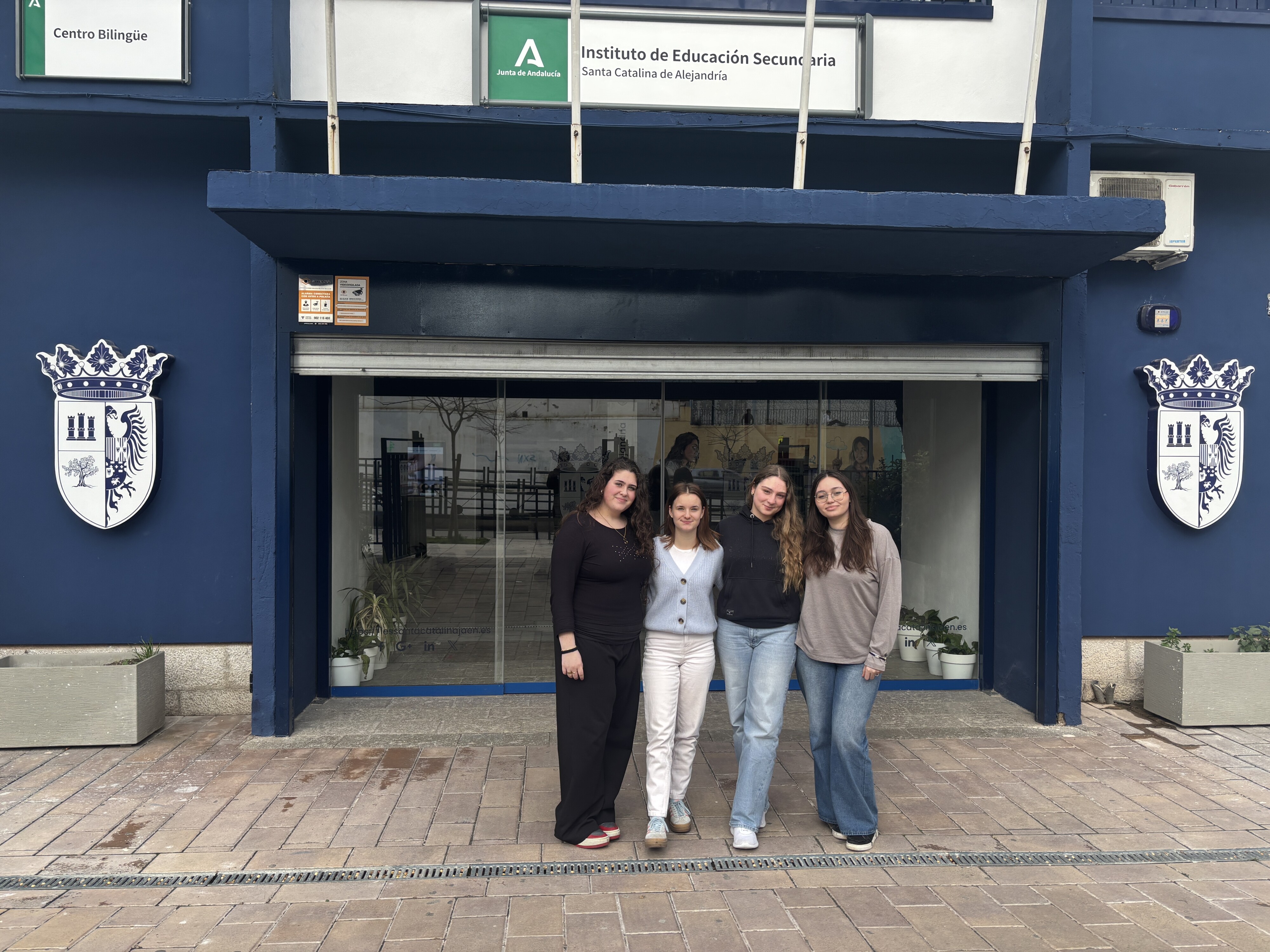 Vier Schülerinnen der Gesamtschule stehen vor dem Eingang der Partnerschule I.E.S. Santa Catalina de Alejandría in Jaén. Über dem Eingang ist der Schulname auf einem Schild zu sehen.