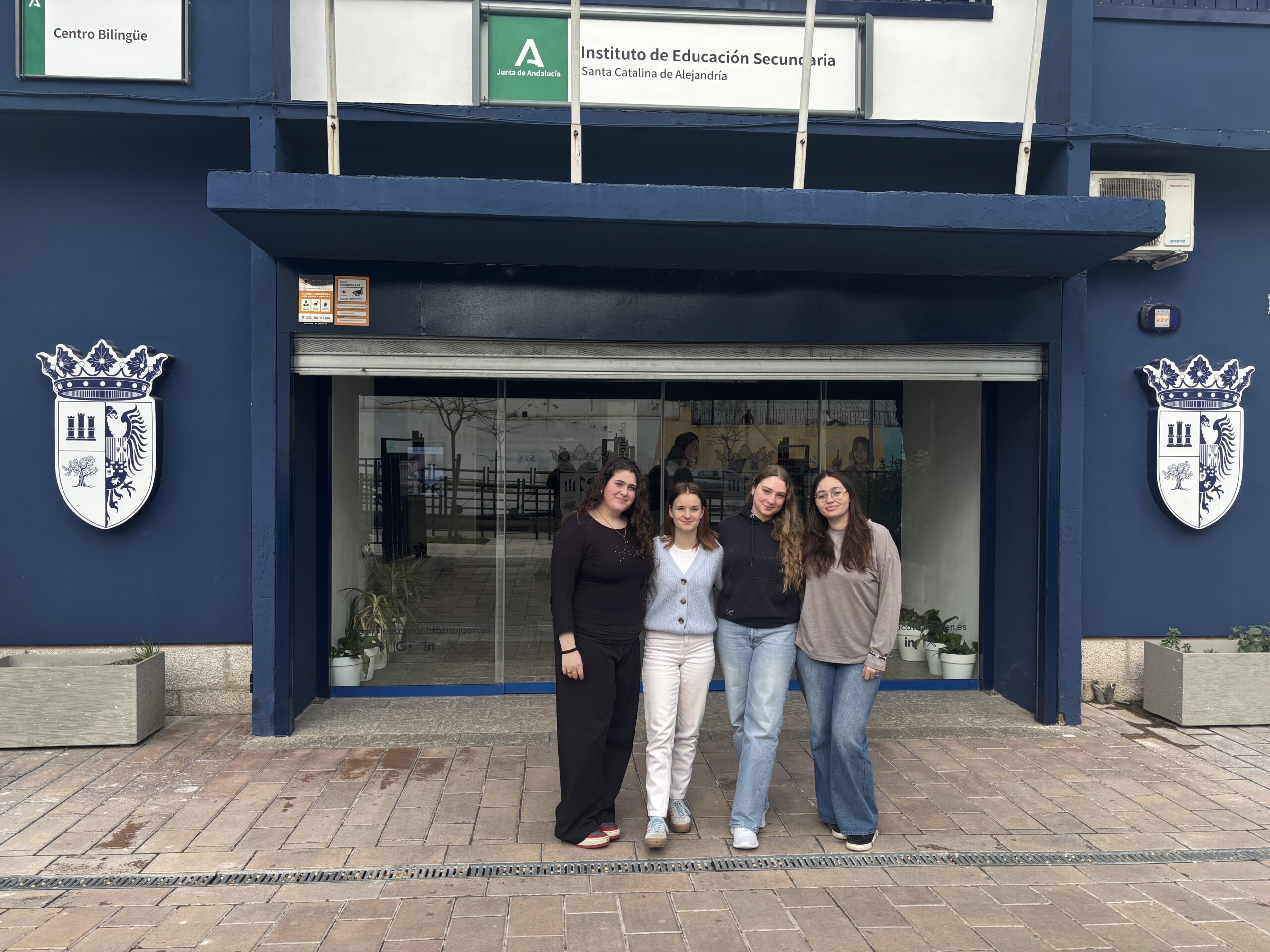 Vier Schülerinnen der Gesamtschule stehen vor dem Eingang der Partnerschule I.E.S. Santa Catalina de Alejandría in Jaén. Über dem Eingang ist der Schulname auf einem Schild zu sehen.