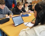 Mehrere Schüler:innen sitzen in Gruppen an zusammengestellten Tischen und arbeiten mit Tablets und Arbeitsblättern. Eine Lehrkraft steht daneben und unterstützt eine Gruppe beim Bearbeiten der Aufgaben.