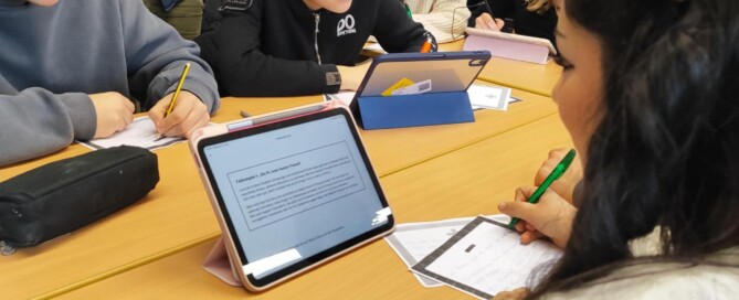 Mehrere Schüler:innen sitzen in Gruppen an zusammengestellten Tischen und arbeiten mit Tablets und Arbeitsblättern. Eine Lehrkraft steht daneben und unterstützt eine Gruppe beim Bearbeiten der Aufgaben.