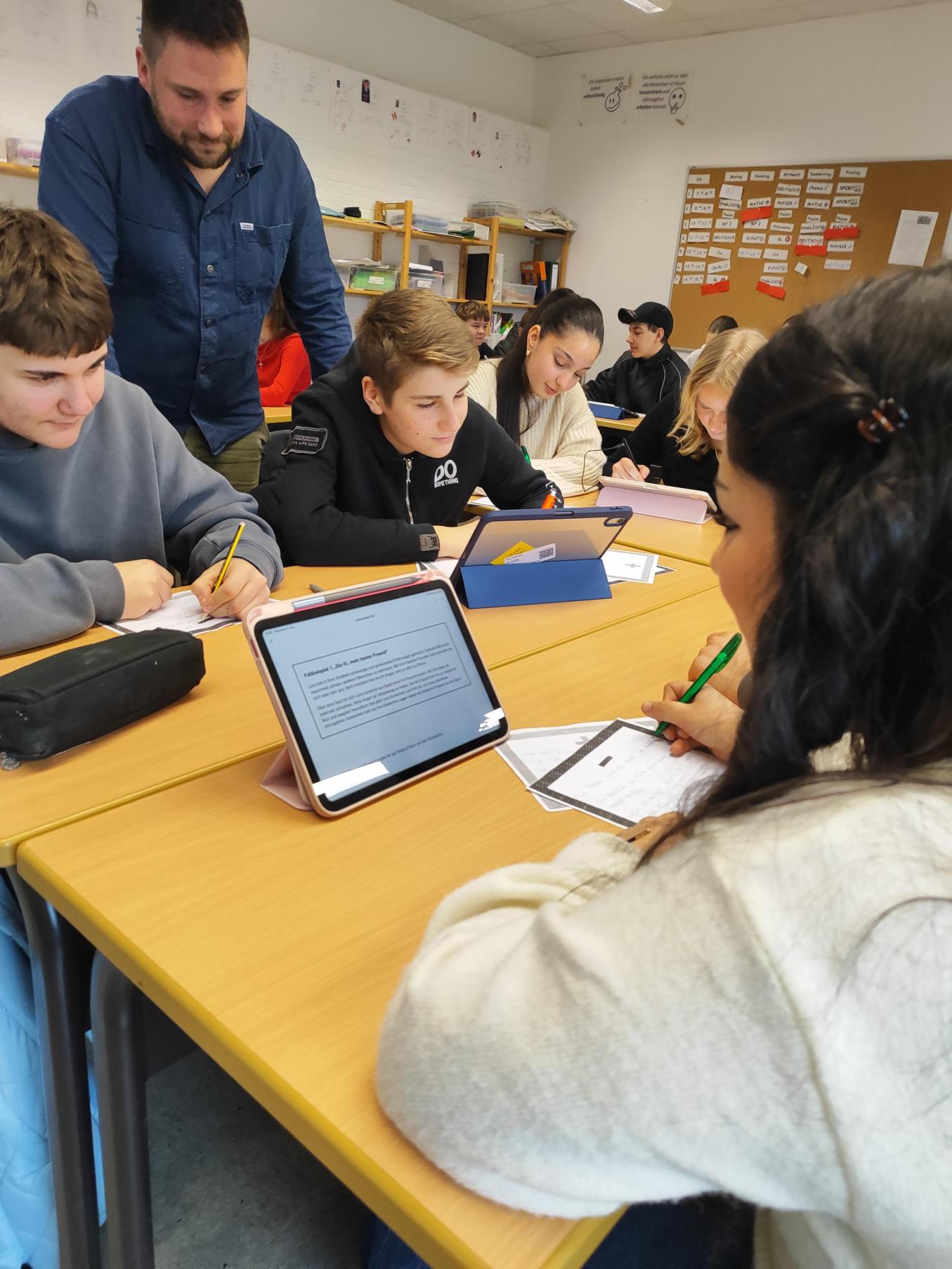 Mehrere Schüler:innen sitzen in Gruppen an zusammengestellten Tischen und arbeiten mit Tablets und Arbeitsblättern. Eine Lehrkraft steht daneben und unterstützt eine Gruppe beim Bearbeiten der Aufgaben.