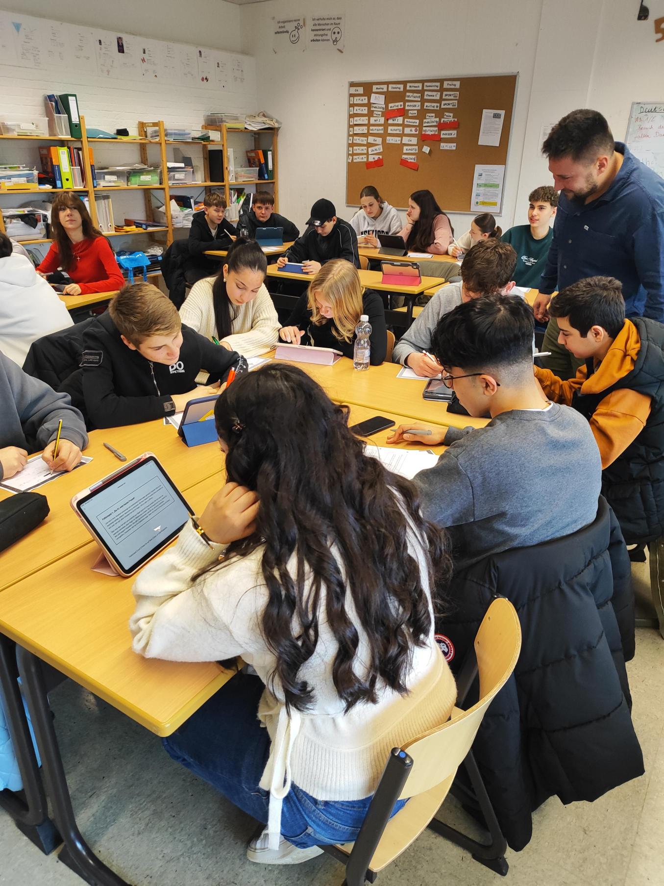 Schüler:innen arbeiten in kleinen Gruppen an Tablets und Papieraufgaben. Eine Lehrkraft steht zwischen den Tischen und begleitet die Arbeit. Der helle Klassenraum ist mit Whiteboard, Pinnwand und Regalen ausgestattet.