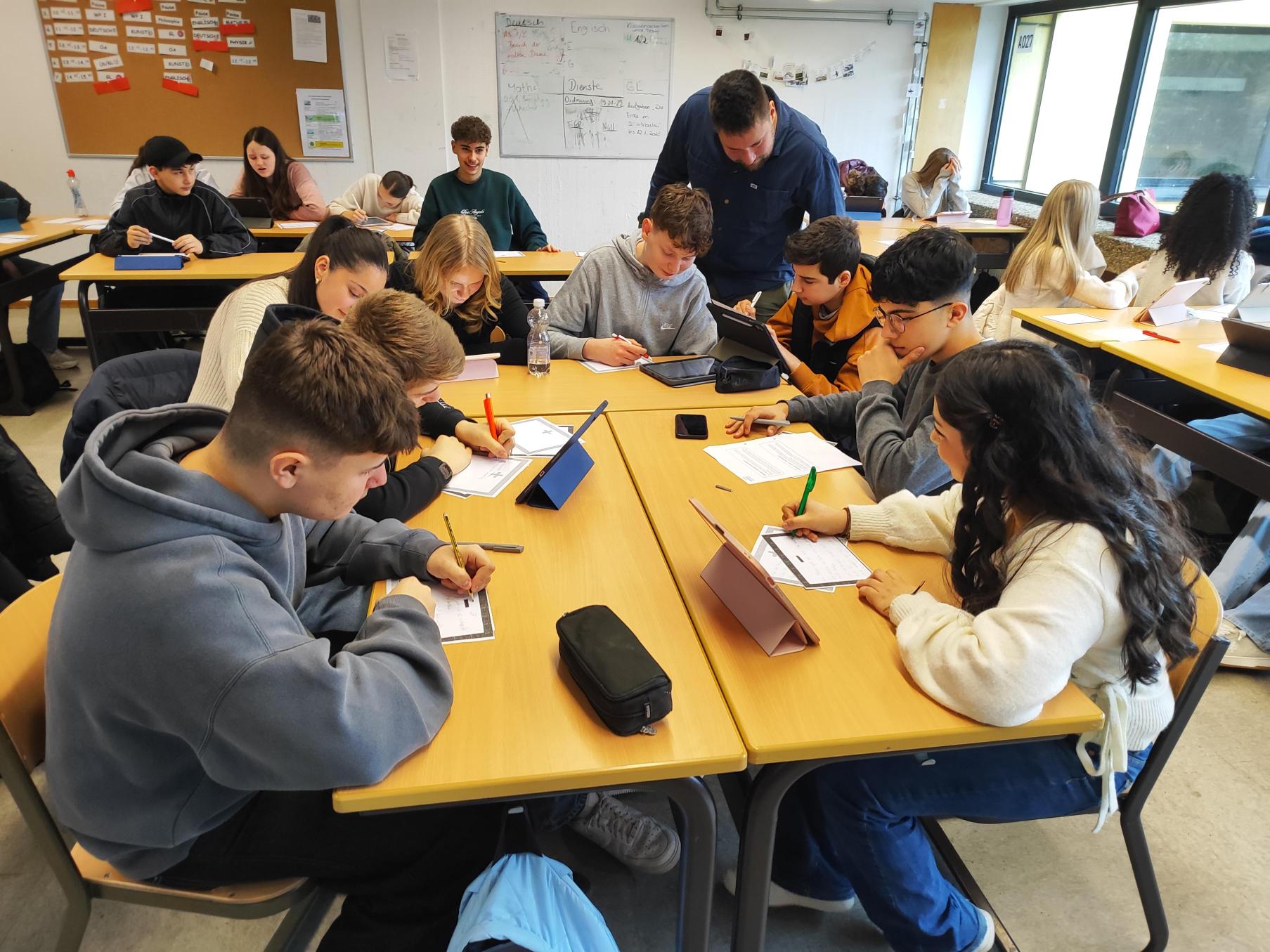 Ein Klassenraum mit mehreren Gruppentischen: Schüler:innen schreiben auf Arbeitsblättern oder nutzen Tablets, während eine Lehrkraft sich zu einer Tischgruppe beugt und beratend hilft. Im Hintergrund sind weitere Lernende und ein Pinnboard zu sehen.