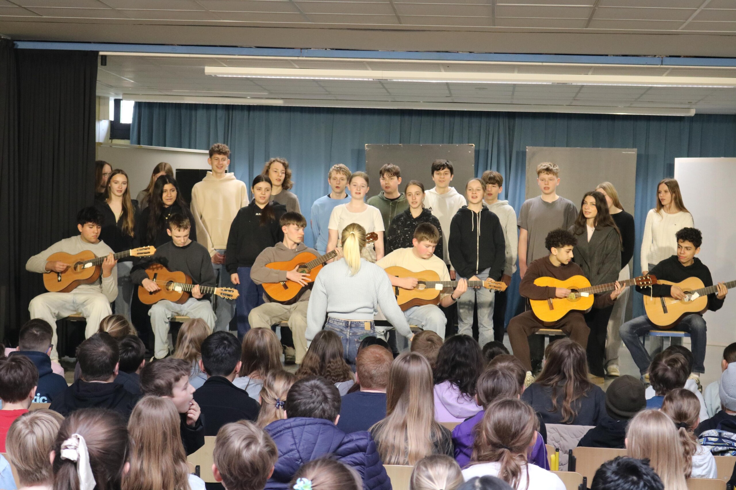 Die Klasse 8a präsentiert einen musikalischen Beitrag mit Gesang und Gitarren vor einem großen Publikum in der Aula.