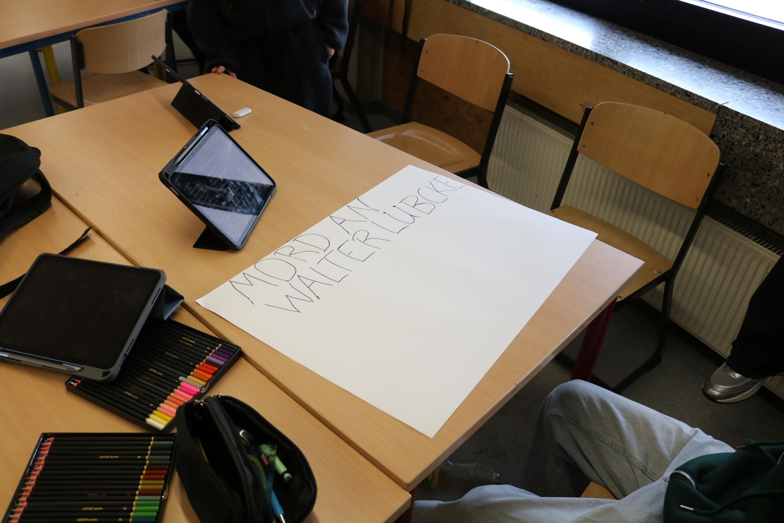 Plakat mit der Aufschrift „Mord an Walter Lübcke“ liegt auf einem Tisch, daneben Tablets und Stifte zur Vorbereitung eines Projekts.