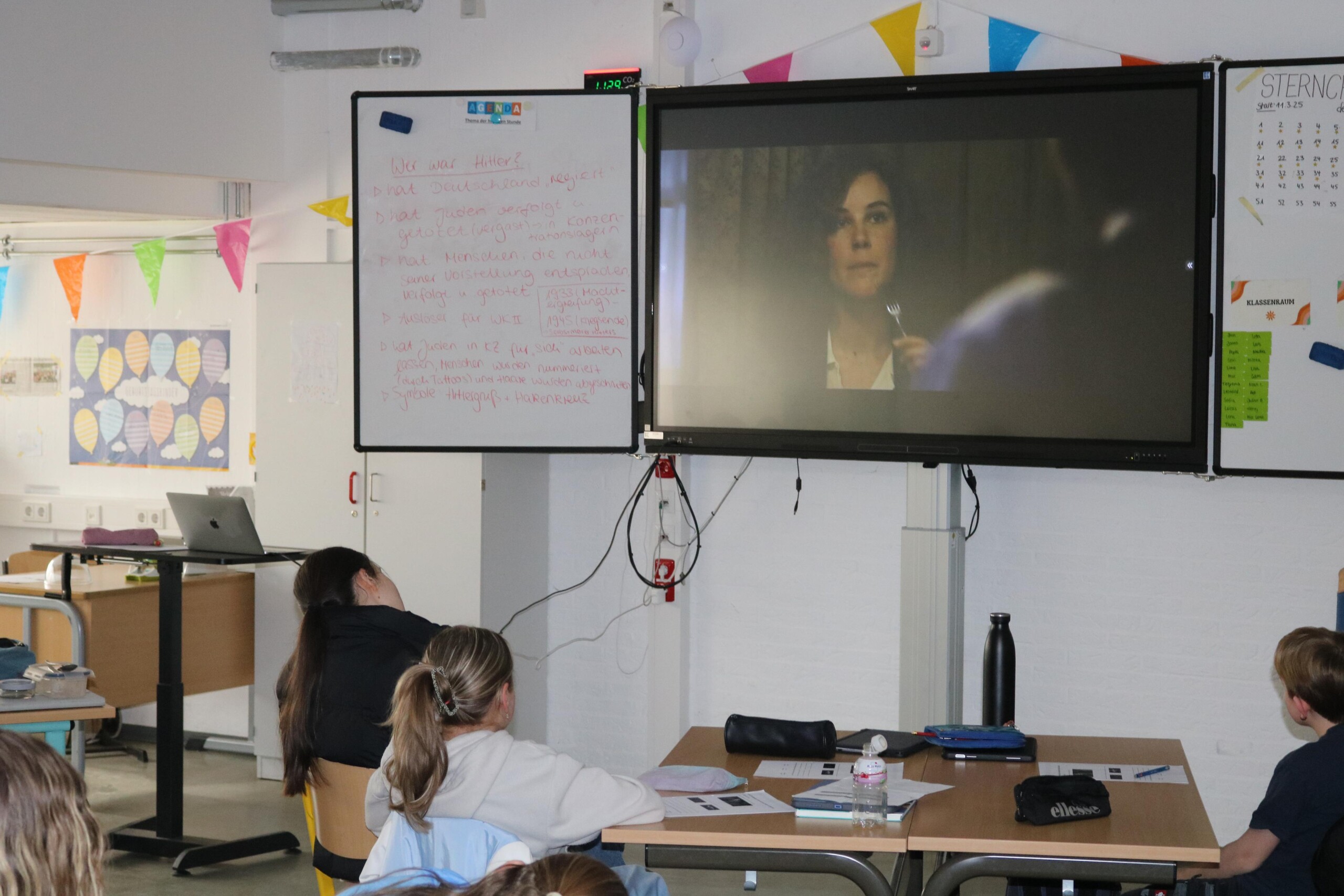 Schüler:innen schauen im Klassenraum eine Filmszene auf einem Bildschirm und arbeiten parallel mit Unterrichtsmaterialien.