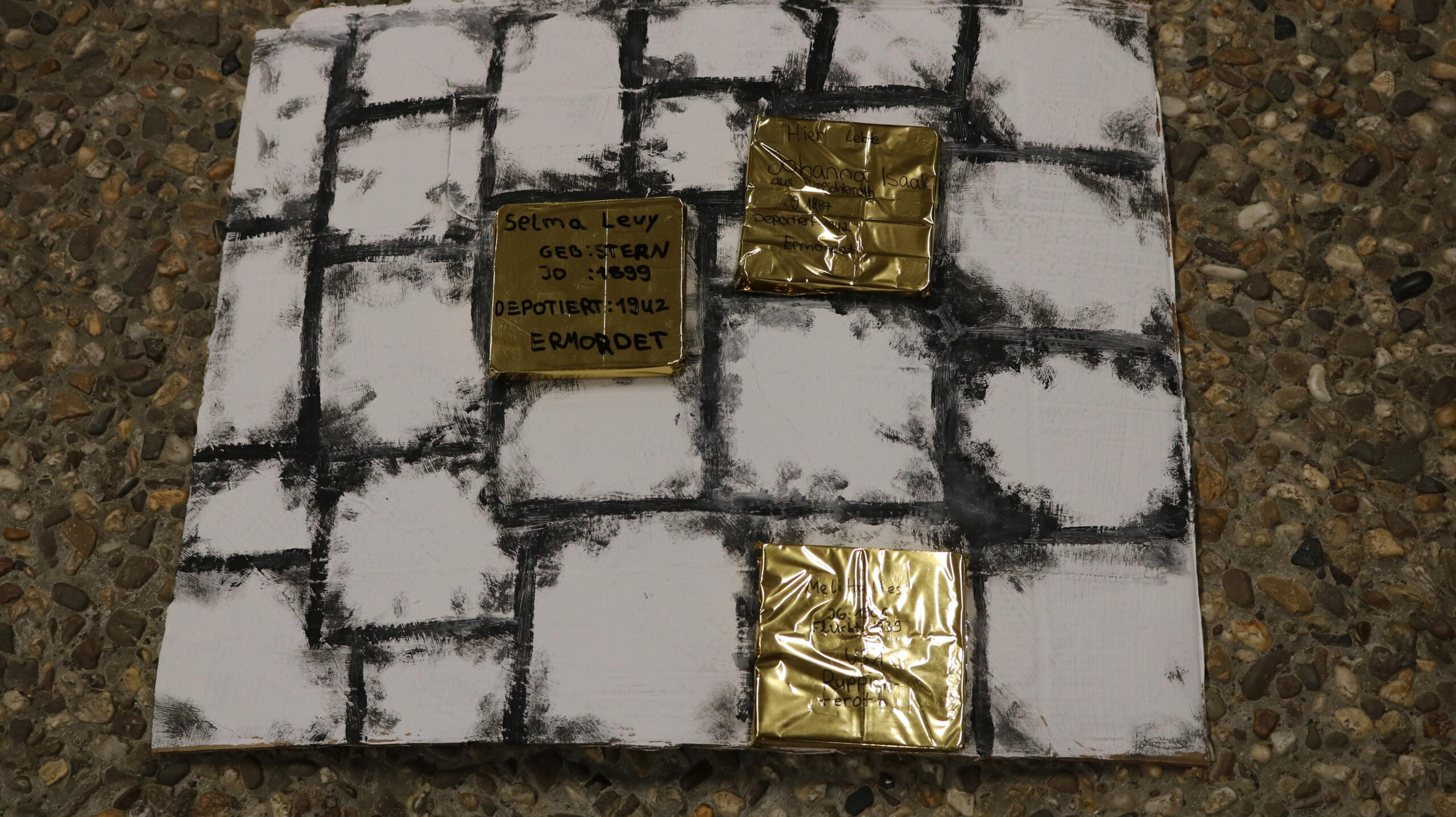 Gebastelte Nachbildung von Stolpersteinen auf einem bemalten Untergrund mit aufgemalten Pflastersteinen, beschriftet mit Namen und Daten von NS-Opfern.