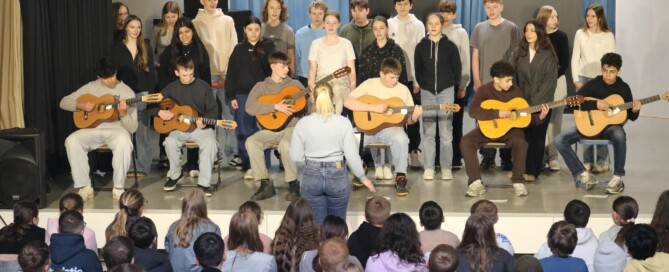 Schüler:innen der Klasse 8a stehen singend auf der Bühne, begleitet von mehreren Gitarrenspielern.