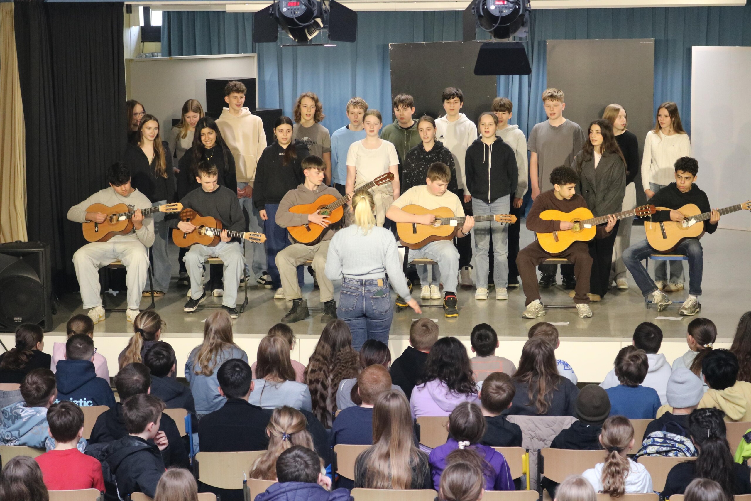 Schüler:innen der Klasse 8a stehen singend auf der Bühne, begleitet von mehreren Gitarrenspielern.
