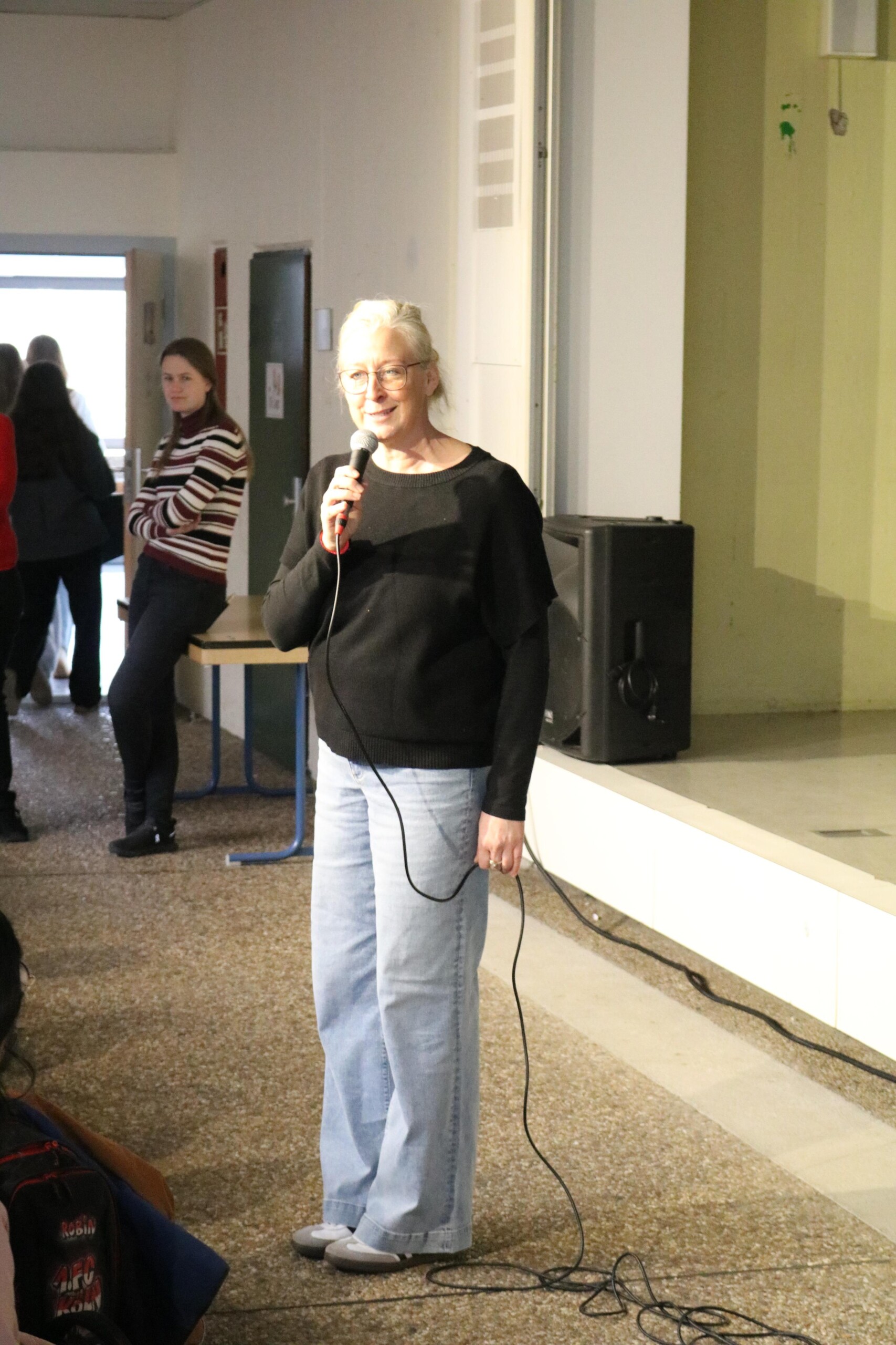 Lehrerin Christine Jahn steht mit Mikrofon vor der Bühne der Aula und begrüßt das Publikum zur Open Stage.