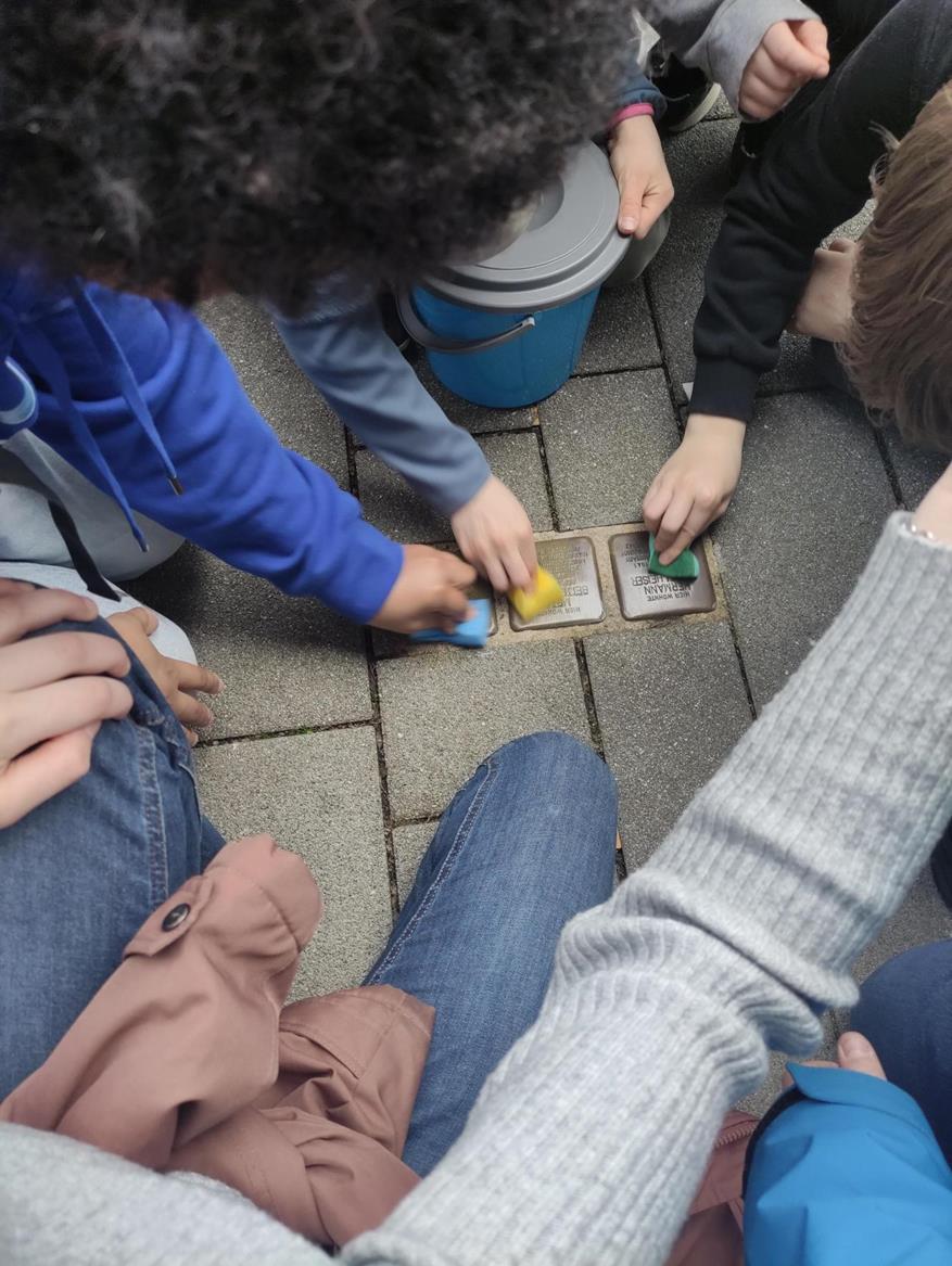 Schüler:innen reinigen mit Schwämmen mehrere Stolpersteine im Gehweg, daneben steht ein Eimer mit Wasser.