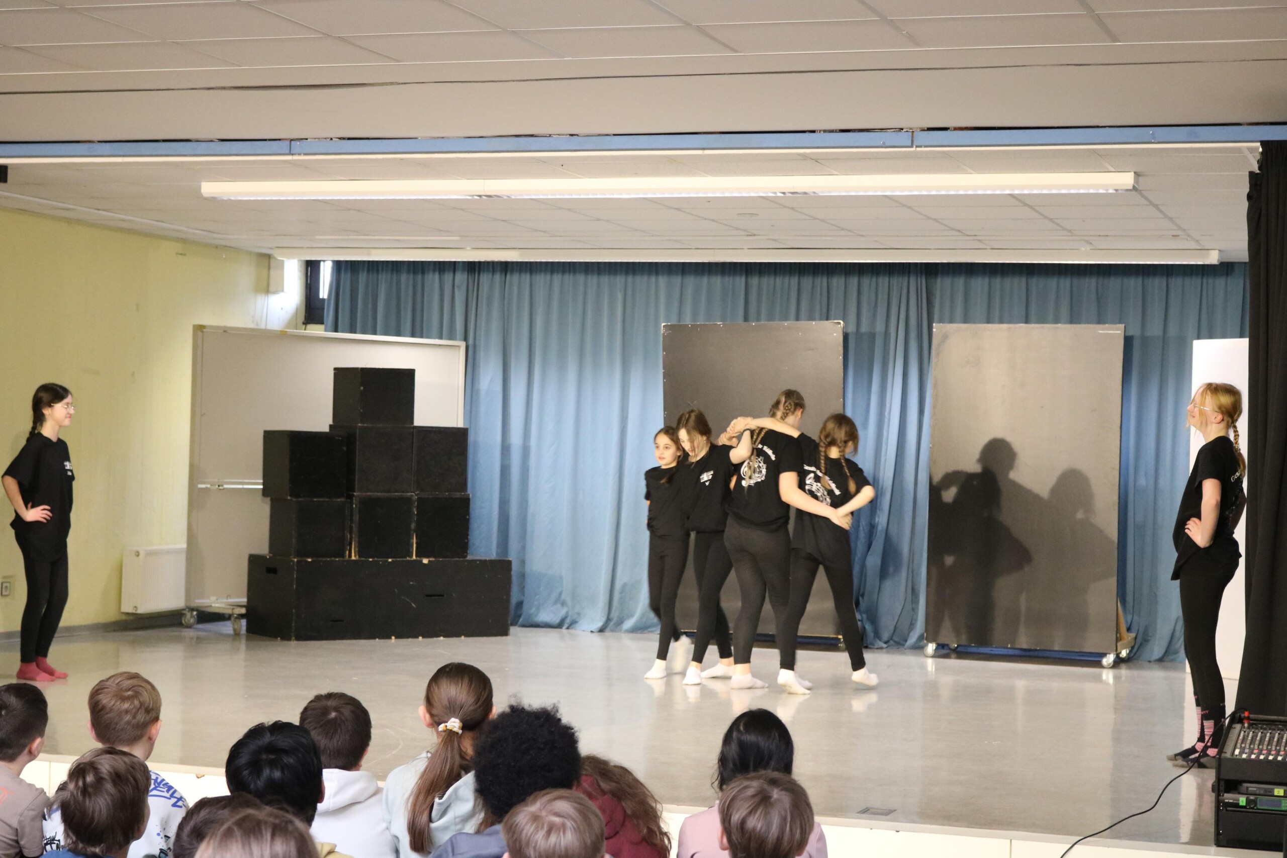 Schüler:innen der Klasse 6d führen auf der Bühne eine Choreografie in schwarzen Outfits vor, während das Publikum vor der Bühne zuschaut.