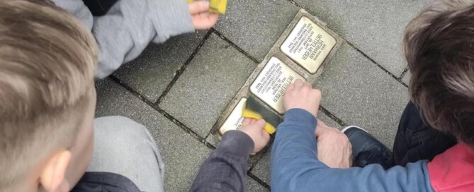 Schüler:innen knien um mehrere Stolpersteine auf dem Gehweg und säubern diese gemeinsam mit Schwämmen.