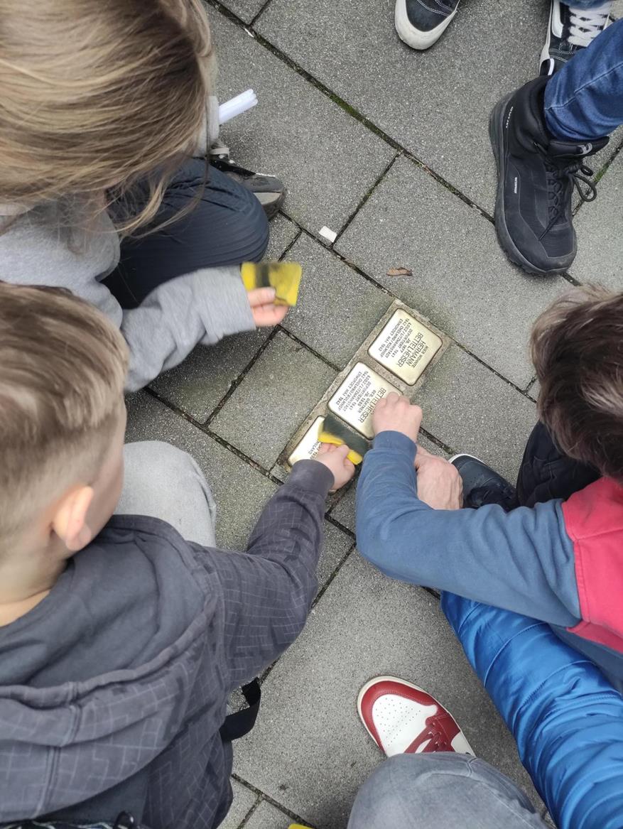 Schüler:innen knien um mehrere Stolpersteine auf dem Gehweg und säubern diese gemeinsam mit Schwämmen.