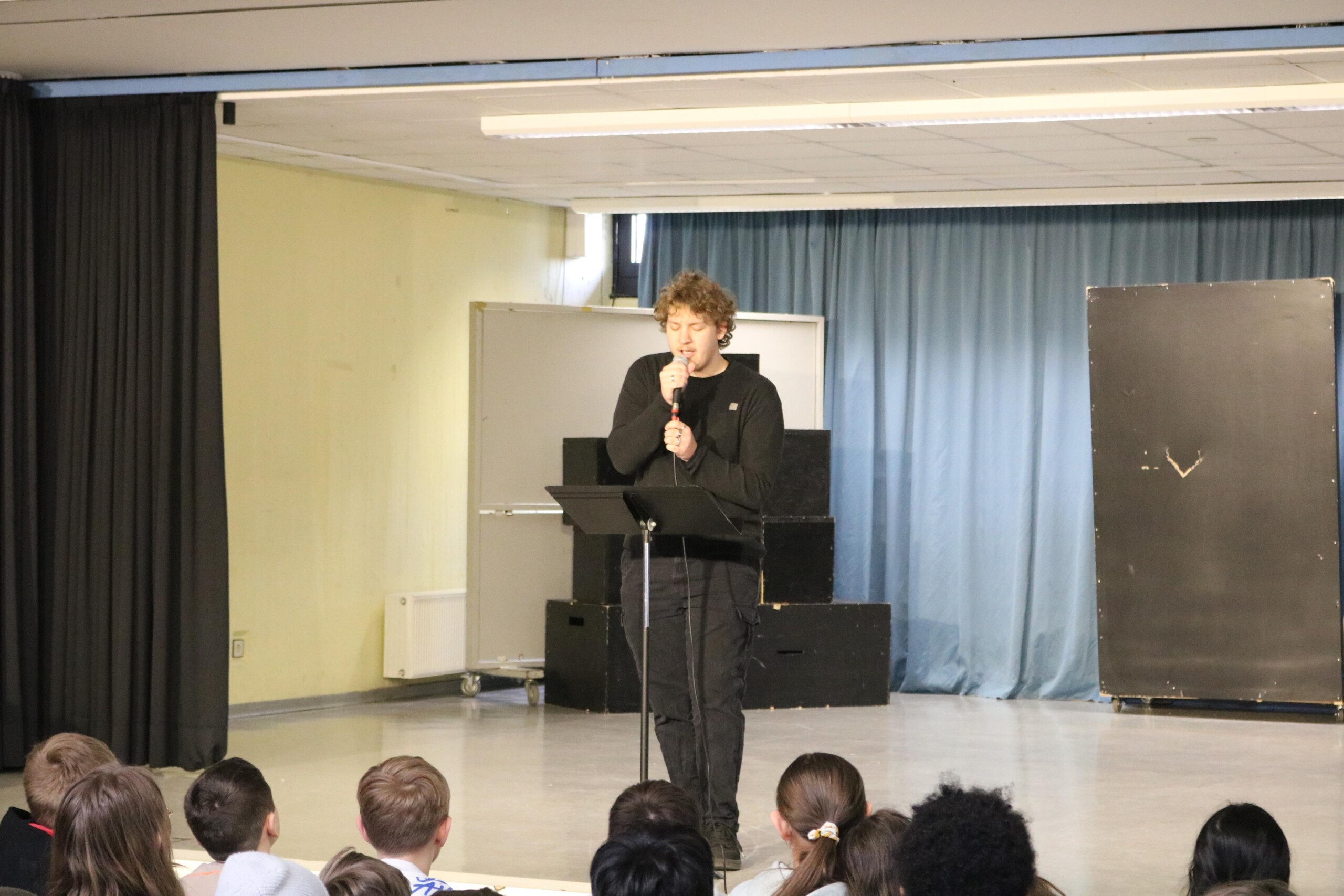 Finn Kalsner aus der Q2 singt an einem Mikrofon auf der Bühne der Aula während das Publikum aufmerksam zuhört.