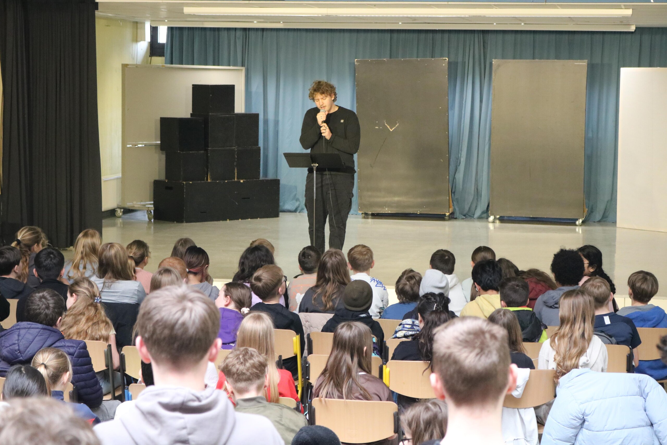 Blick aus dem Publikum auf Finn Kalsner, der auf der Bühne singt, während viele Schüler:innen im Saal zuhören.
