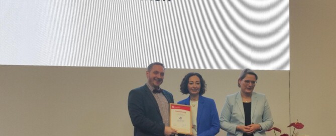 Auf einer Bühne wird die Auszeichnung „Verbraucherschule Silber“ an die Gesamtschule Waldbröl überreicht. Im Hintergrund ist eine große Präsentationsfläche mit dem Namen der Schule zu sehen.